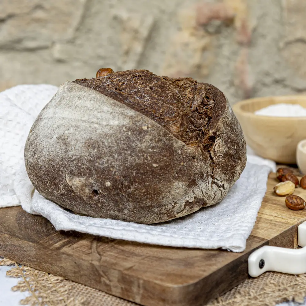 Pan artesanal de algarroba sin gluten con avellanas enteras, sobre tabla de madera y paño blanco, ideal para dietas saludables