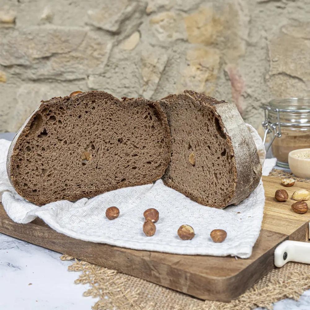 Pan de algarroba sin gluten partido por la mitad, con miga oscura y trozos de avellanas visibles, sobre tabla de madera y fondo rústico