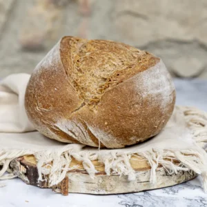 Pan de avena sin gluten de corteza rústica y color tostado, sobre una base de madera con un paño beige de flecos, fondo de piedra natural