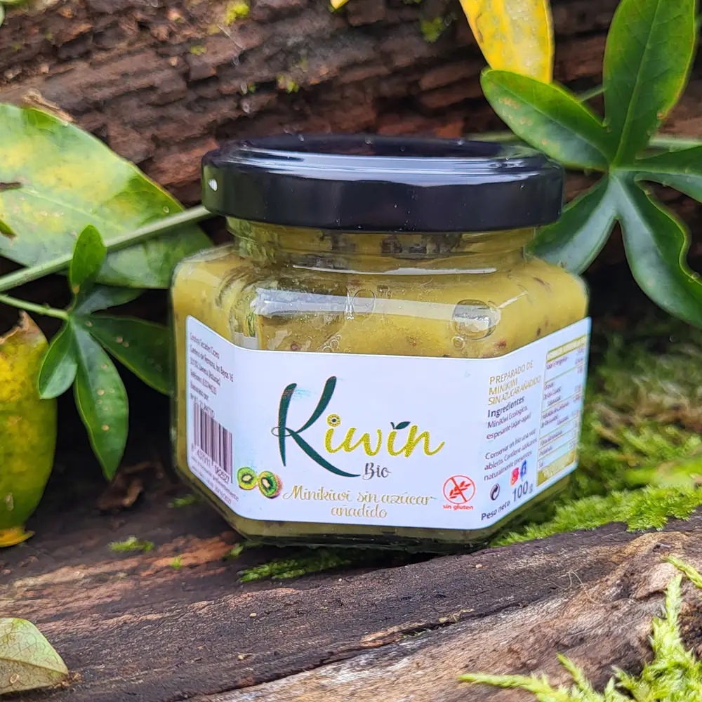 Tarro de mermelada de kiwi orgánica, con etiqueta que muestra el logo de la marca 'Kiwiin Bio' y la descripción 'Mermelada sin azúcar añadido'. El tarro está rodeado de hojas verdes y otros elementos naturales, sobre un fondo de madera oscura.