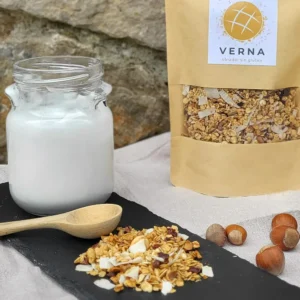 Granola artesanal sin gluten de Verna con avellanas, coco y nibs de cacao, presentada junto a yogur en tarro de cristal y envase ecológico del obrador Verna. Desayuno saludable y nutritivo.