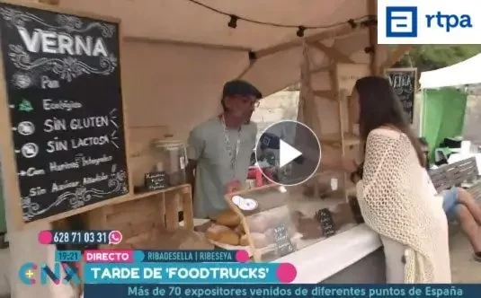 El vendedor de Verna Sin Gluten responde a las preguntas de la reportera de Conexión Asturias (RTPA) durante el festival Fartukarte en Ribadesella. En el stand se muestran panes artesanos, ecológicos, sin gluten y sin lactosa, elaborados por Verna Sin Gluten.